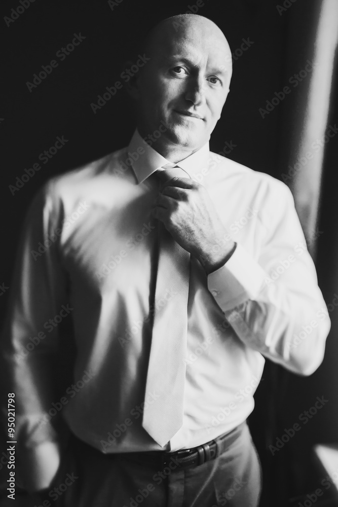 solid bald man in shirt standing near the window straightens tie solid bald man in shirt standing near the window straightens tie