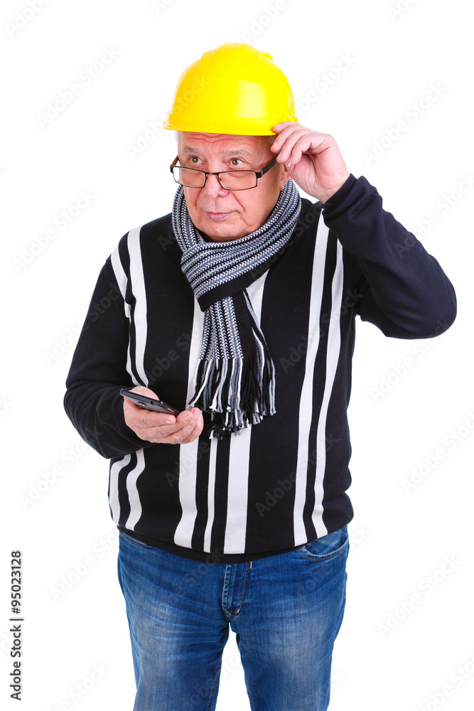 Serious old senior man in sweater, jeans, scarf, glasses, wearing yellow construction hat. Holding cell phone in hand and thinking. Touching helmet, head, glasses. Isolated, plain white background