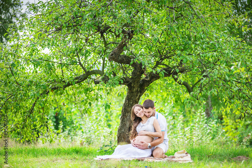 Happy young couple expecting baby, pregnant woman with husband touching belly, sitting on green grass  © Andrey Bandurenko