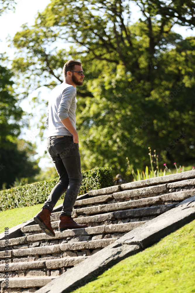 Man wearing jeans,boots and a grey shirt walking up a stone steps outside on a sunny summer day. 