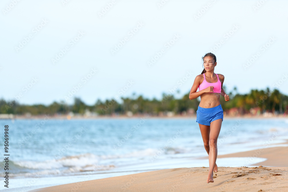 Young fit woman running on beach against sky. Full length of determined female runner in sports clothing. Jogger is exercising at sea shore during sunny day.