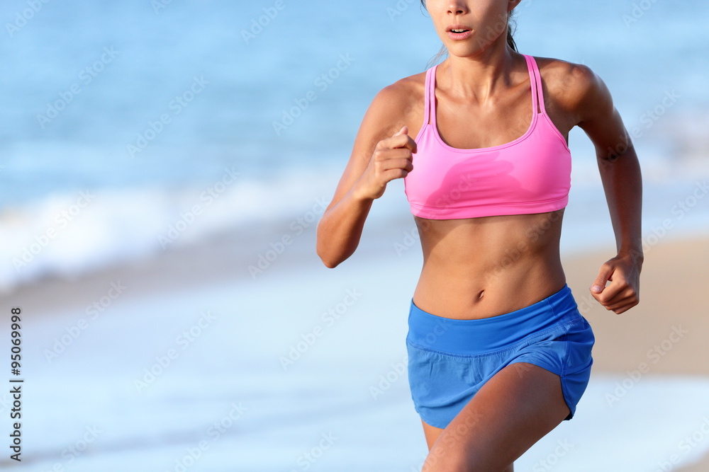 Midsection of determined woman jogging on beach. Young female is wearing  sports bra and shorts. Sporty runner is exercising on sunny day. Stock  Photo