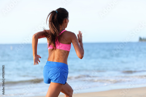Fit young woman jogging on beach against sky. Rear view of determined female is in sports clothing. Runner is exercising at sea shore during sunny day.