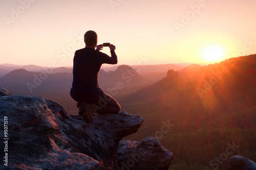 Young man in black sportswear is sitting on sharp cliff and taking photos by smart phone
