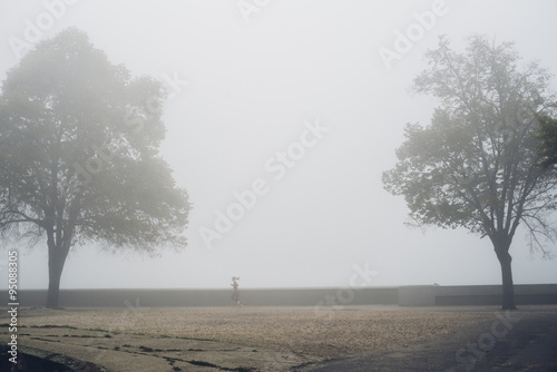 Young lady jogging on the Tisza river embankment on foggy November morning with two big trees framing the pictire photo