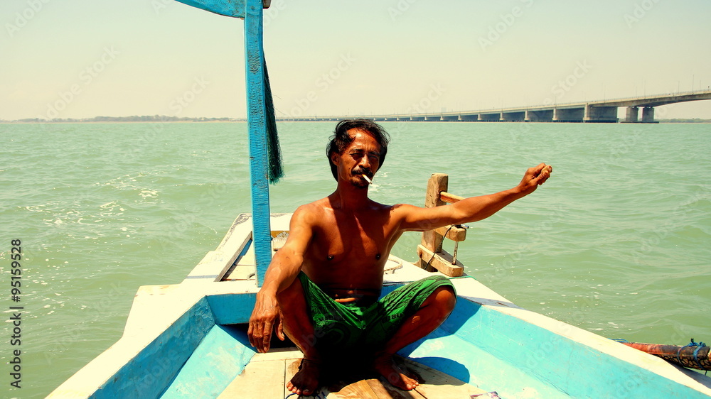 braungebrannter indonesischer Fischer steuert rauchend sein Boot vor der Brücke Suramadu