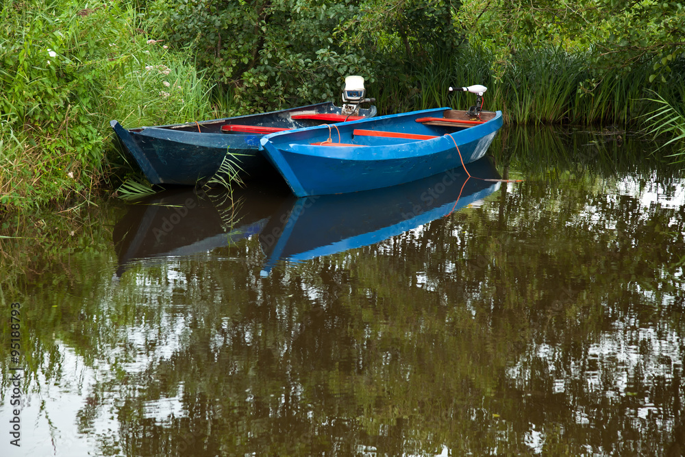 Two blue boats parked on a lake surrounded with plants and bushes. Two blue boats parked on a lake surrounded with plants and bushes.
