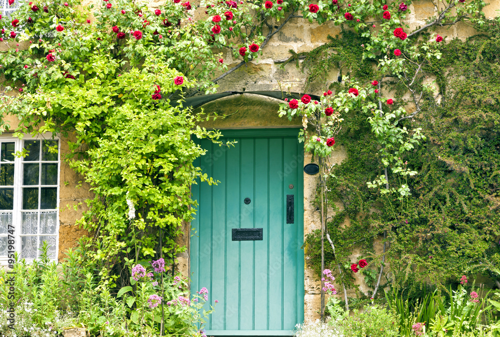 Naklejka premium Green wooden doors in an old traditional English stone cottage surrounded by climbing red roses and flowers
