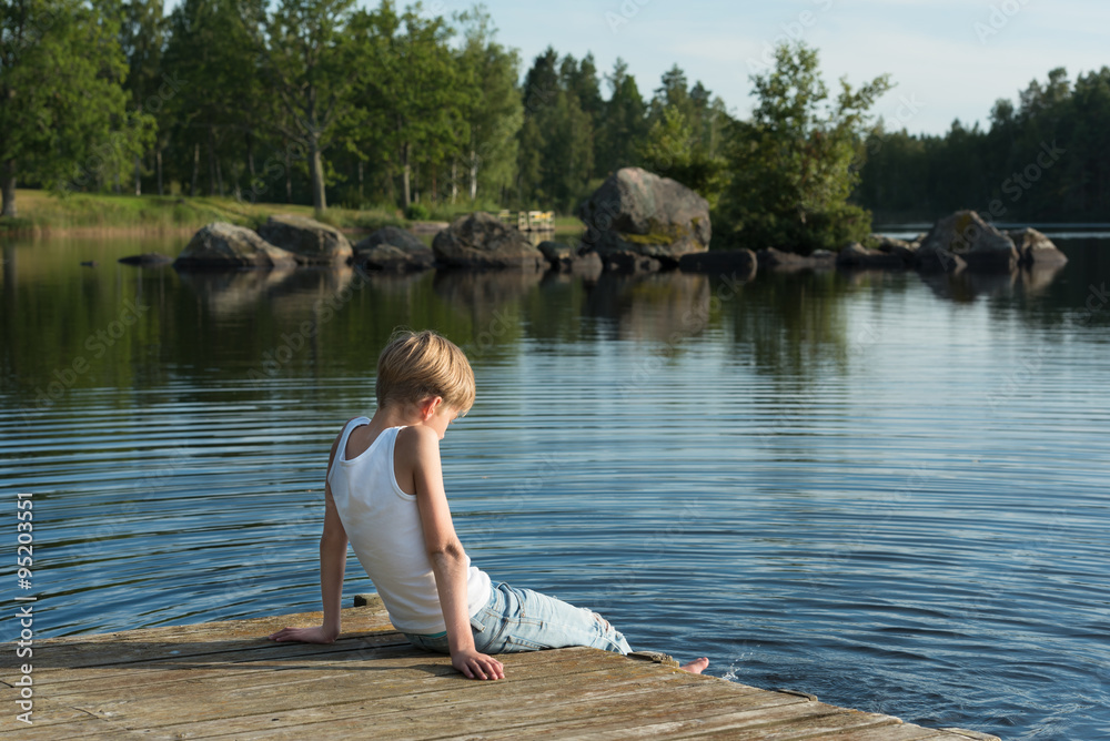 Fototapeta premium Boy relaxing on a jetty. He's sitting in the sunshine and looking out over the beautiful lake Skiren in the swedish countryside.