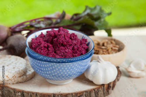 Beetroot pesto in a blue bowl on a wooden table with garlic, beetroot and rice cakes on the back, selective focus