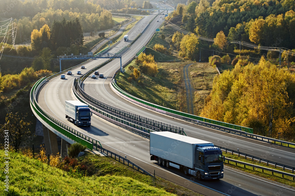 Asphalt highway with electronic toll gates in autumn woodland. Three trucks on the road. The bridge spanning the valley. View from above. Sunny day with bright fall colors.