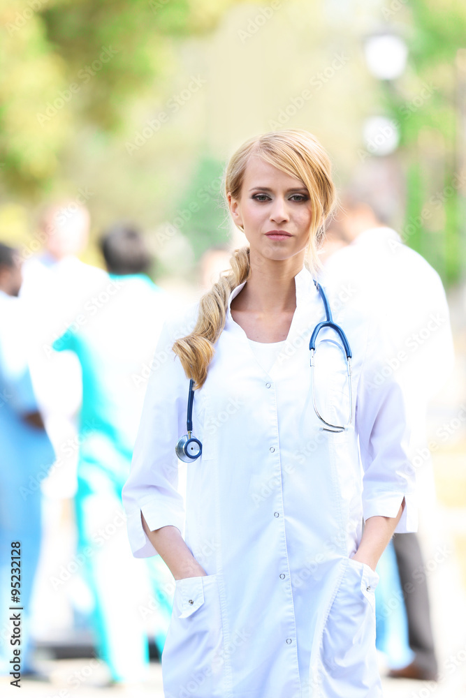 Beautiful smart woman doctor with clipboard in hands standing against blurred group of medics Beautiful smart woman doctor with clipboard in hands standing against blurred group of medics