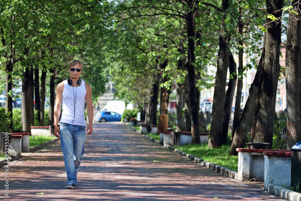 Obraz premium Man in sunglasses goes on alley with green trees and benches
