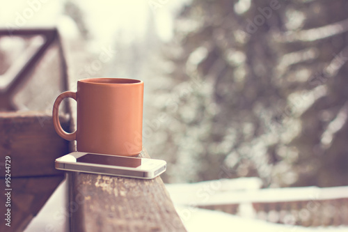 A cup with a hot drink and mobile phone on the background of the winter forest