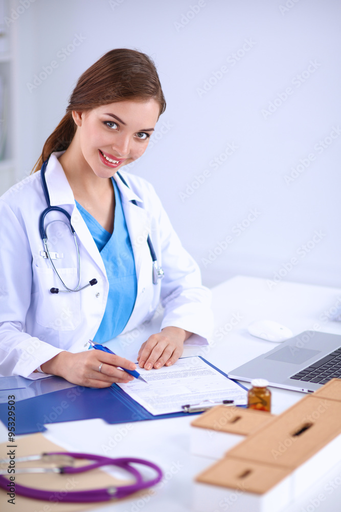 Beautiful young smiling female doctor sitting at the desk and writing Beautiful young smiling female doctor sitting at the desk and writing