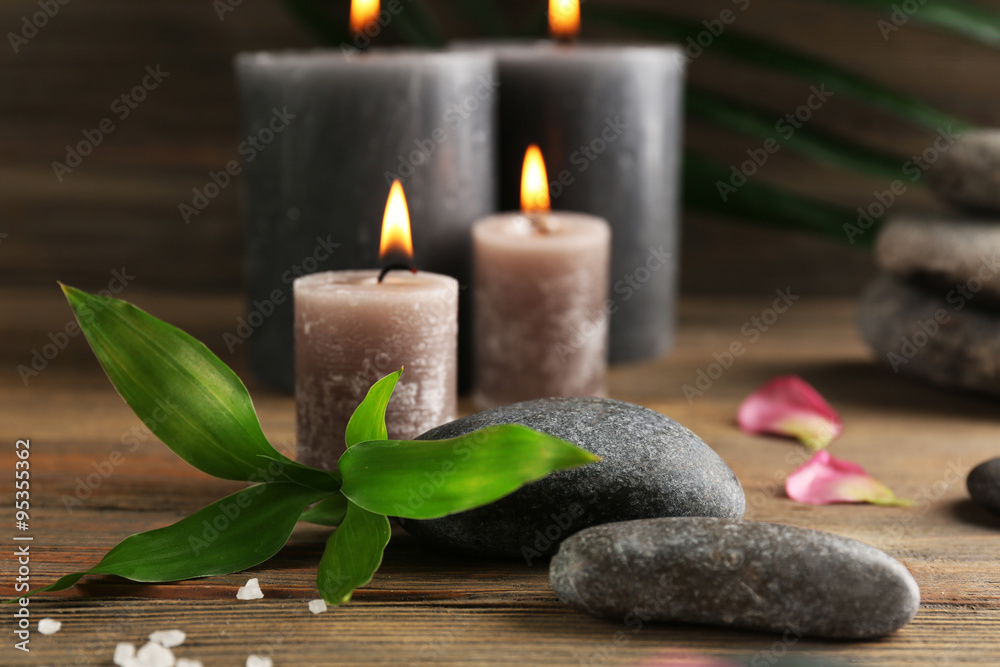 Relax set contains alight wax candles with flowers and pebbles on wooden background, focus on green leaf Relax set contains alight wax candles with flowers and pebbles on wooden background, focus on green leaf