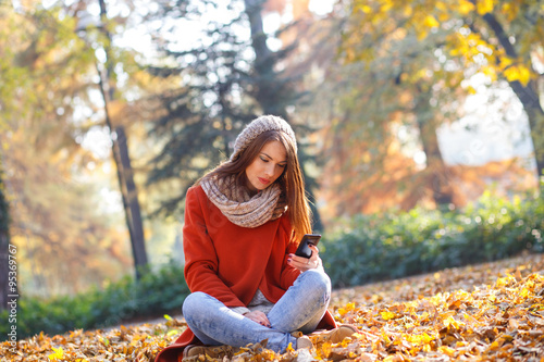 Young brunette woman with smart phone relaxing in park in autumn. She looking at phone and texting.