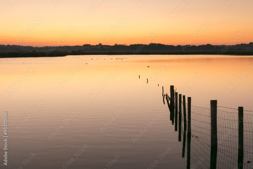 Orange and yellow sunset over still water lake with wooden fence posts to right Orange and yellow sunset over still water lake with wooden fence posts to right