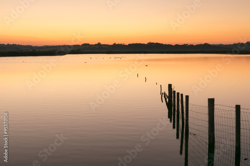 Orange and yellow sunset over still water lake with wooden fence posts to right