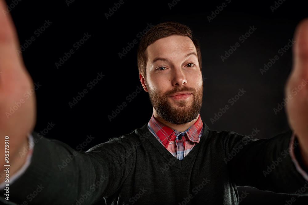 Life style concept. Young man with a beard in shirt and pullover holding mobile phone and making photo of himself while standing against black background. Life style concept. Young man with a beard in shirt and pullover holding mobile phone and making photo of himself while standing against black background.