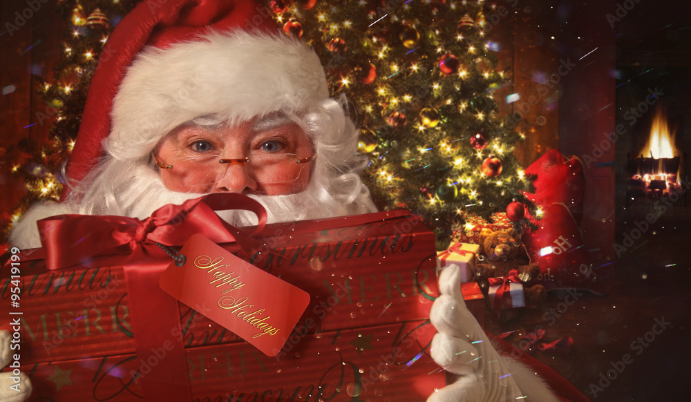 Closeup of Santa holding gift with Christmas scene in background - obrazy, fototapety, plakaty 