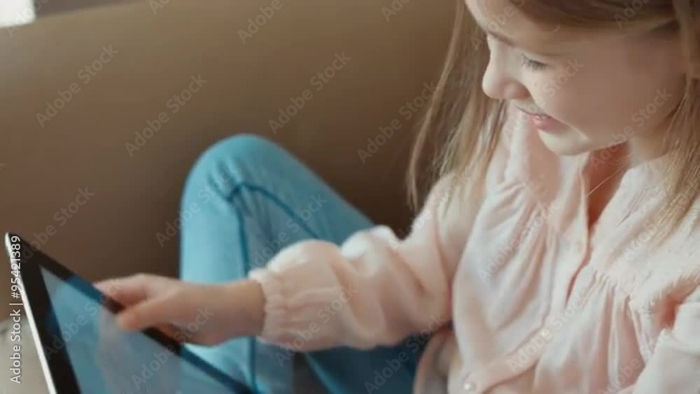 Closeup Blond little girl is online with tablet pc and sitting on the couch. Child laughing and looking at camera Closeup Blond little girl is online with tablet pc and sitting on the couch. Child laughing and looking at camera