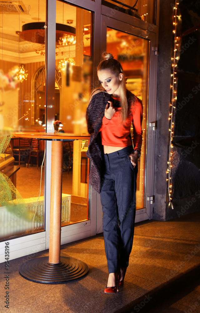 Beautiful young woman poses near bright cafe on the street at night