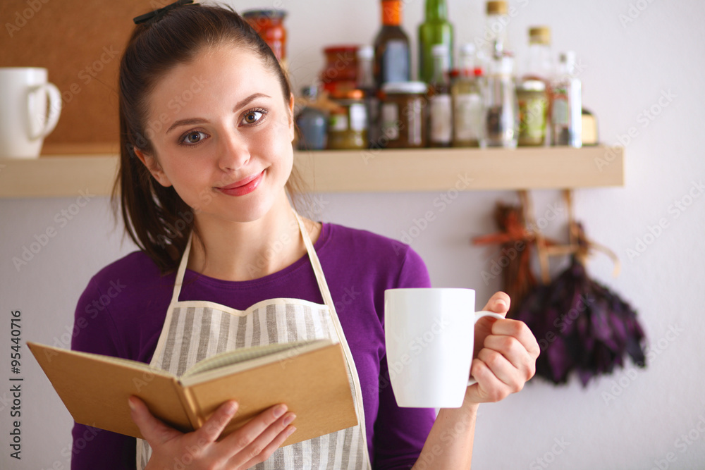 A young woman standing in her kitchen drinking tea and holding a cookbook A young woman standing in her kitchen drinking tea and holding a cookbook