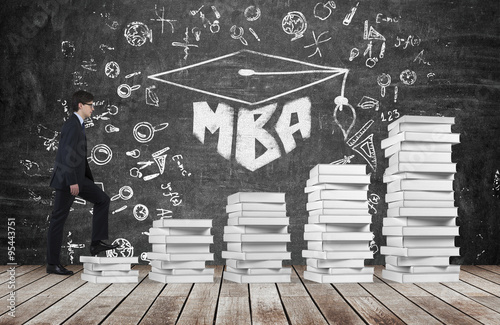A man is going up using a stairs which are made of white books to reach graduation hat. The written word MBA is drawn on the black chalkboard which symbolises a professional business education.