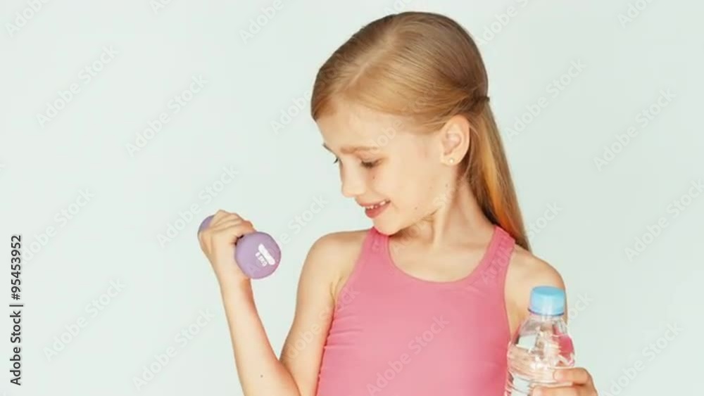 Child turns around at camera. Girl holding dumbbell and bottle of water on a white background and laughing. Zooming Child turns around at camera. Girl holding dumbbell and bottle of water on a white background and laughing. Zooming