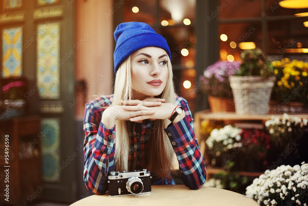 Obraz premium Portrait of beautiful stylish young woman sitting in street cafe and drinking coffee. Hipster with old retro camera. Model looking aside. Lights and flowers background. City lifestyle. Toned