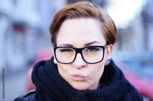 Fröhliche Frau mit heller, reiner Haut und kurzem Haar, die eine moderne Brille und ein Schal trägt und mit Kussmund in die Kamera lächelt. Nette kurzhaarige Frau mit langen Wimpern. photo