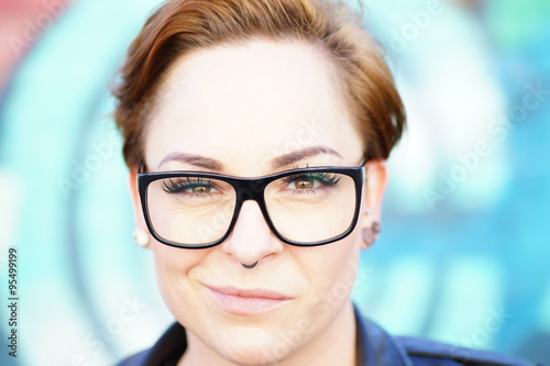 Fotografie Pfiffige Frau mit heller, reiner Haut und kurzem Haar, die eine schwarze, moderne Brille trägt und mit lächelnden Mund frontal in die Kamera schaut