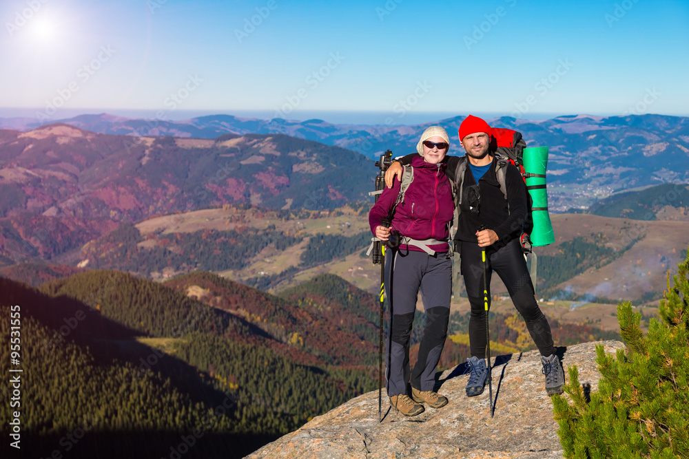 Male and Female Hikers with Backpacks Sporty Clothing and Trekking Poles Staying on High Rock with Mountain View with Autumnal Colors Forest and Blue Sky Shining Sun on Background