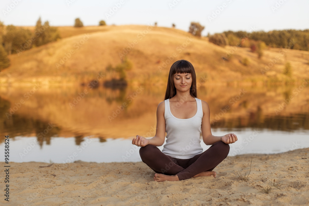 Young woman doing yoga in the lotus position at the shore of lake. Relax in nature.