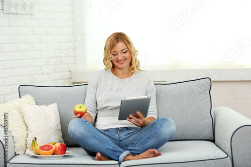 Beautiful woman with fresh fruits and digital tablet on home interior background