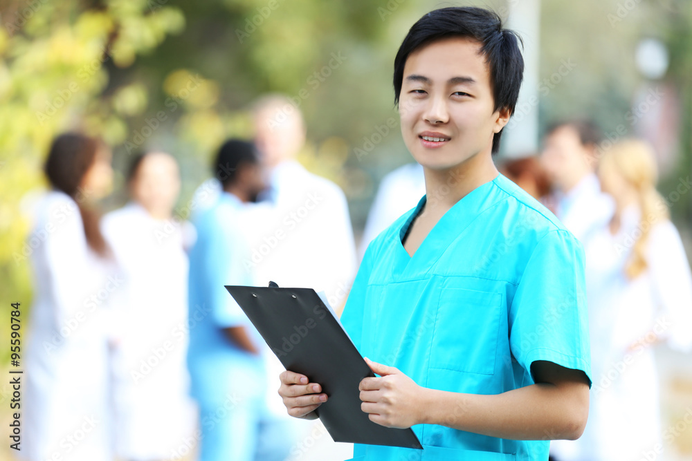 Young smiling doctor with clipboard in hands standing against unfocused group of medical workers Young smiling doctor with clipboard in hands standing against unfocused group of medical workers