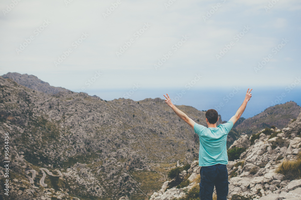 Young man standing on cliff with hands raised on top of mountain and looking to the hills of Sa Colobra, Majorca, Spain Young man standing on cliff with hands raised on top of mountain and looking to the hills of Sa Colobra, Majorca, Spain