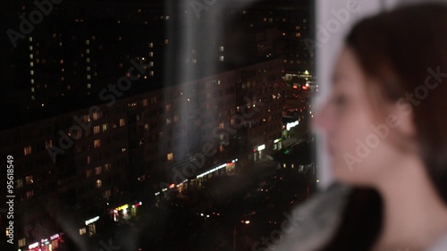 Sad woman sits on window sill, look at night city photo