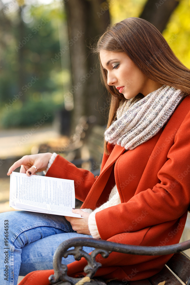 Obraz premium Young girl sitting on a park bench and reading a book, on a beautiful autumn day