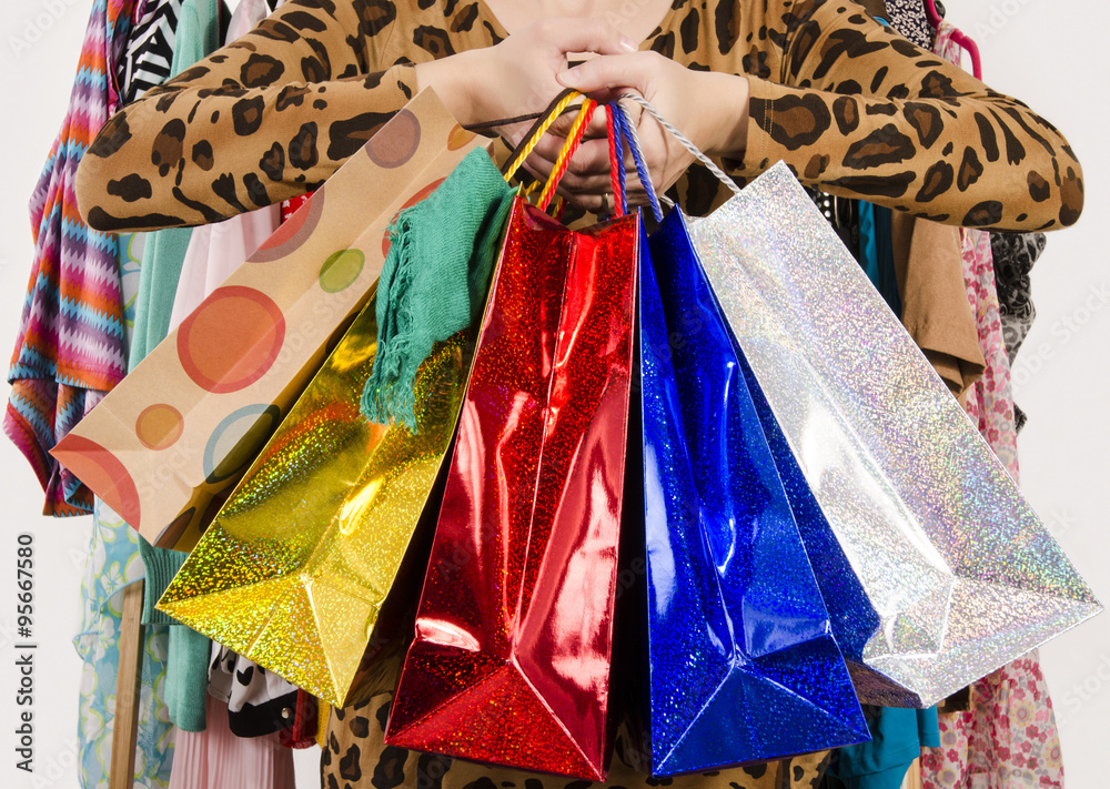 Close up on female hands holding many shopping bags. Unrecognizable woman holding colorful shopping bags with clothes hanged in the background. Girl shopping.