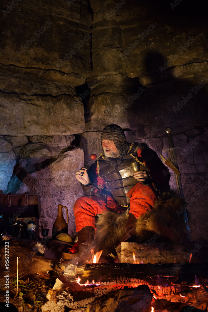 Old medieval King in armor with dish is sitting on furs near the Old medieval King in armor with dish is sitting on furs near the