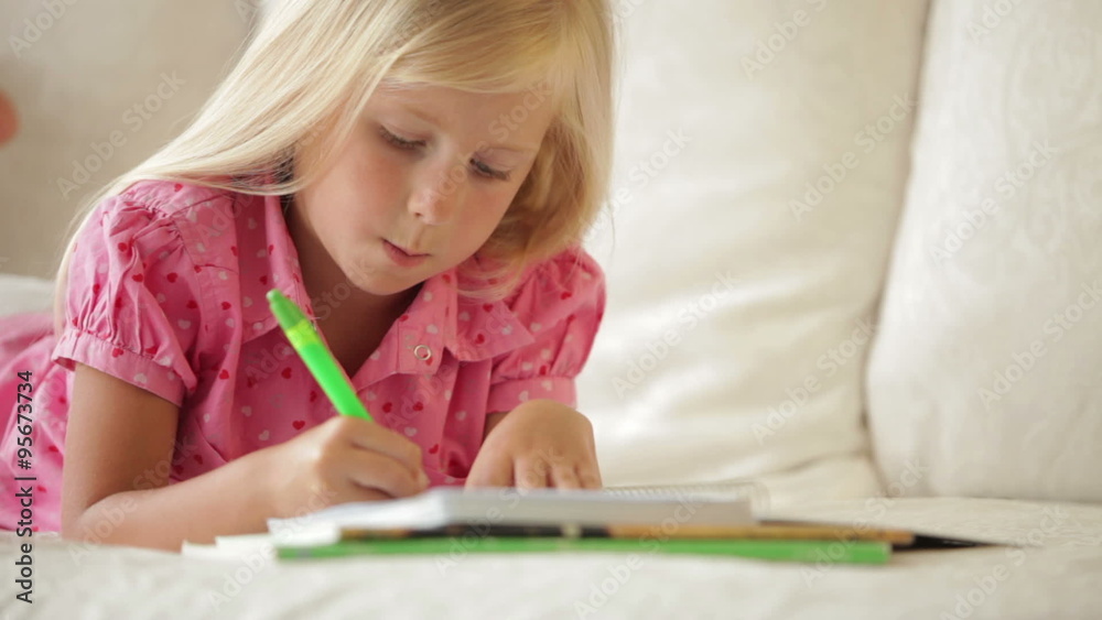 Cheerful little girl lying on sofa writing in notebook and smiling Cheerful little girl lying on sofa writing in notebook and smiling
