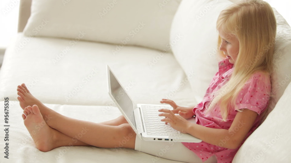 Pretty little girl sitting on sofa using laptop and smiling at camera Pretty little girl sitting on sofa using laptop and smiling at camera