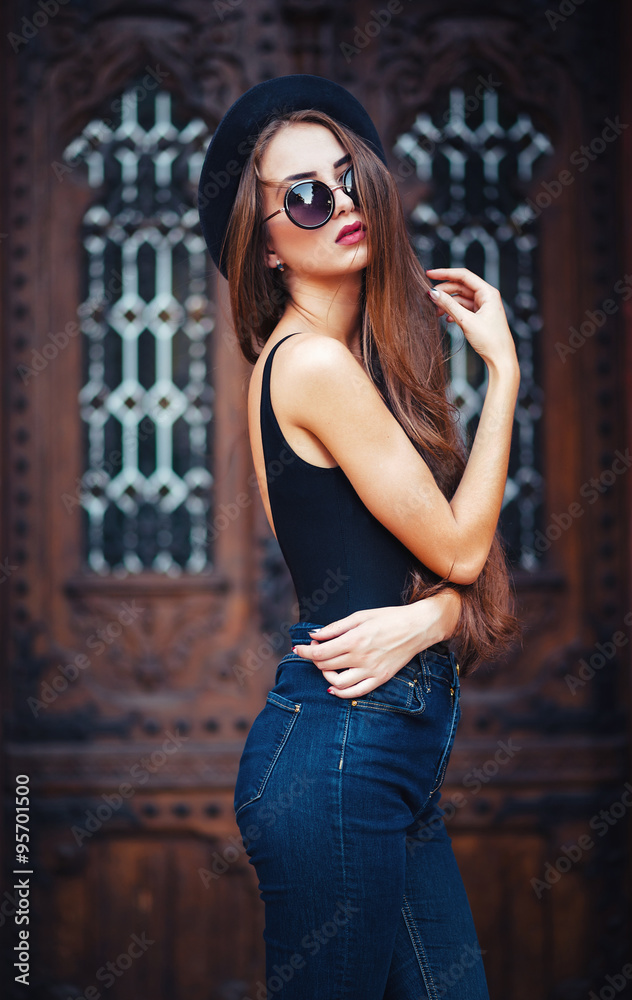 Naklejka premium young beautiful brunette girl in a hat, sunglasses, t-shirt and jeans, with a film camera walks in the old city, holding hand hat for fear of losing it, vertical picture