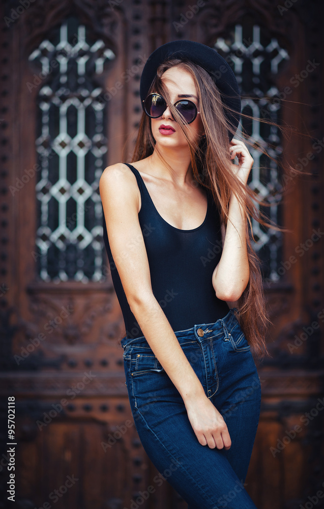 Naklejka premium young beautiful brunette girl in a hat, sunglasses, t-shirt and jeans, with a film camera walks in the old city, holding hand hat for fear of losing it, vertical picture