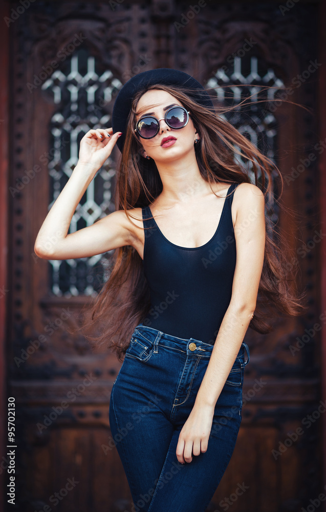 Fototapeta premium young beautiful brunette girl in a hat, sunglasses, t-shirt and jeans, with a film camera walks in the old city, holding hand hat for fear of losing it, vertical picture