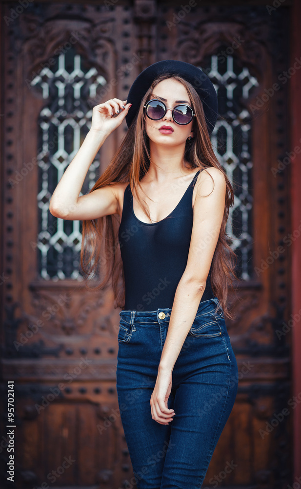 Fototapeta premium young beautiful brunette girl in a hat, sunglasses, t-shirt and jeans, with a film camera walks in the old city, holding hand hat for fear of losing it, vertical picture