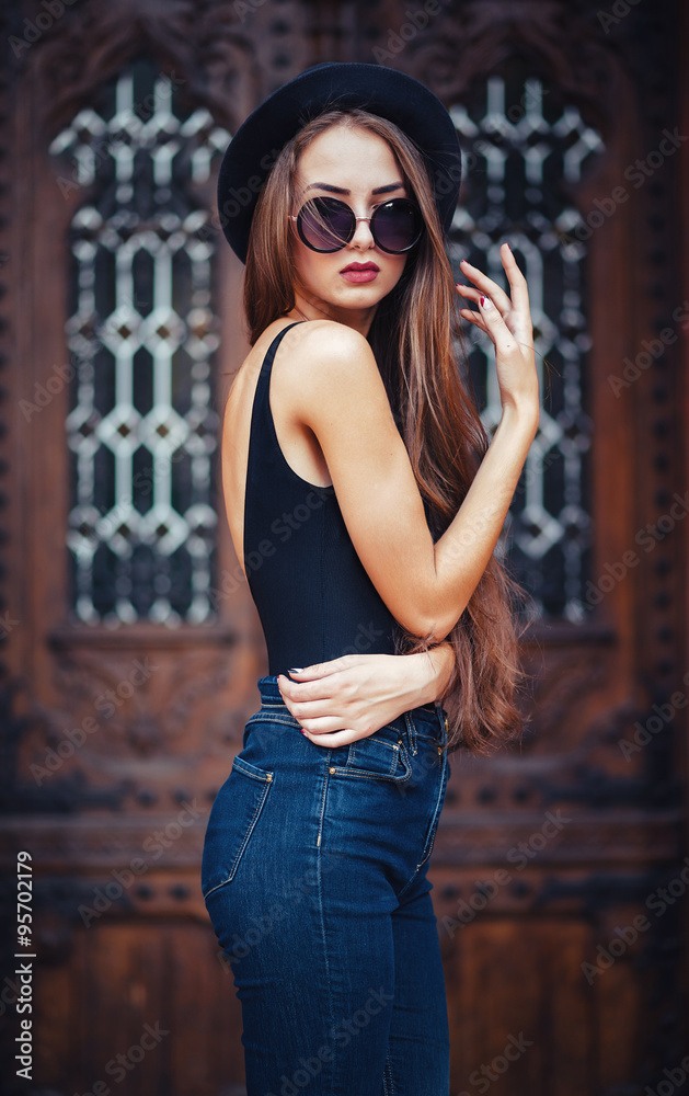 Naklejka premium young beautiful brunette girl in a hat, sunglasses, t-shirt and jeans, with a film camera walks in the old city, holding hand hat for fear of losing it, vertical picture