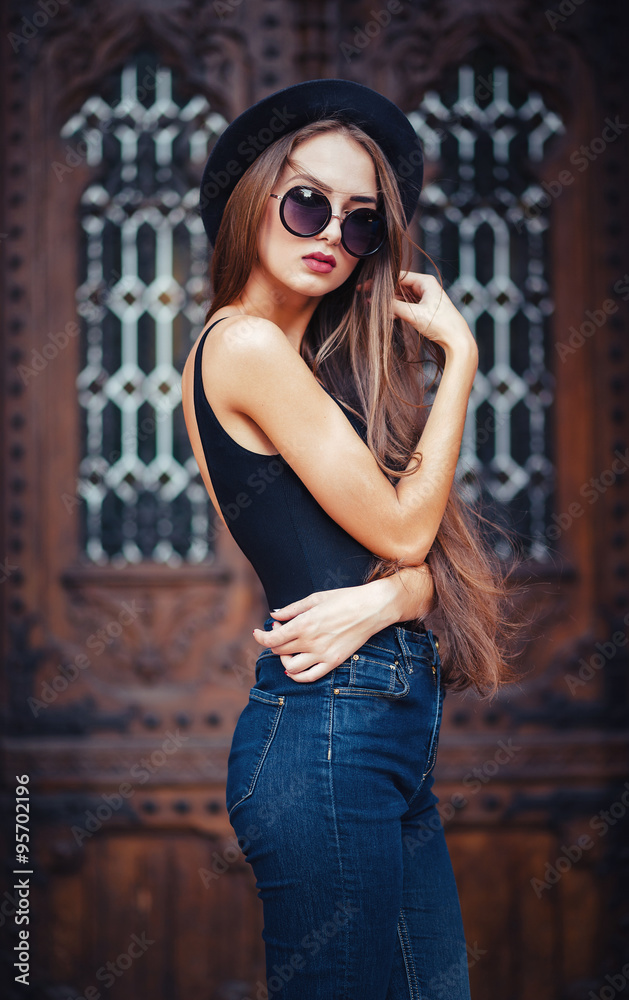 Naklejka premium young beautiful brunette girl in a hat, sunglasses, t-shirt and jeans, with a film camera walks in the old city, holding hand hat for fear of losing it, vertical picture